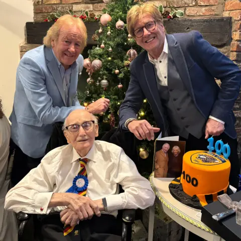 Phillip Norton / BBC A 100-year-old man wearing a birthday rosette and a Hull City tie sits by a table with a birthday cake and a card from the King and Queen. Behind him stand his sons, who are smartly dressed and smiling towards the camera.