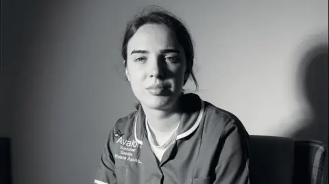 Daisy Moseley Black and white portrait of Summer Woodley. Her hair is tied up and she is wearing a care uniform with the Avalon logo on the chest. She is looking directly at the camera. 