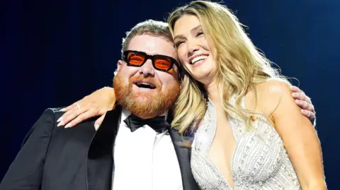 PA Media A smiling Cammy Barnes, who is wearing a dinner suit, is hugged by Delta Goodrem, who is wearing a silver sparkly dress. Cammmy has ginger hair and beard and black framed glasses with red lenses. Delta has long blonde hair.
