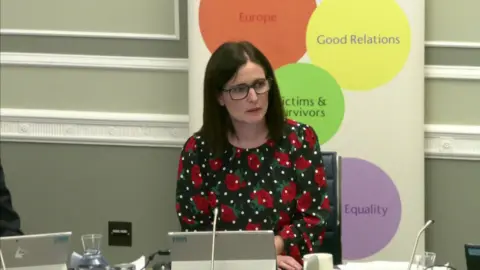 NI Assembly Alliance Party MLA Paula Bradshaw at the Executive Office committee. She has shoulder-length dark hair and is wearing glasses. She is wearing a dark dress with a red rose pattern on it and sitting behind a desk with tablet computer folded in front of her. Behind her is a pull-up banner with words such as "Good Relations" and "Europe" written on coloured circles.