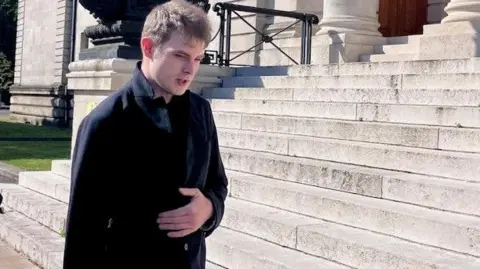 A man with light short hair and a dark coat walks up light stone steps, with his head down.