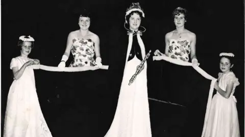 Elizabeth Patterson A black-and-white photograph of three woman and two young girls. The woman in the centre is wearing a black robe and crown, with two women standing either side of her in flowery strapless dresses holding the edges of her robe. At the far ends of both rows stands a young girl wearing white dresses 