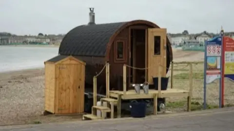 LDRS Beach and sea behind a wooden tube-shaped sauna accessed by wooden steps - the sauna door is open and a small shed stands to its left.