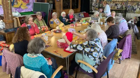 A large group of women seated around several rectangular tables arranged in a square or U‑shaped formation inside a craft or community workshop space. The women are engaged in hands‑on creative activities such as sewing, knitting, or assembling fabric items. Many of them are holding pieces of colourful cloth, thread, or tools. The tables are covered with various crafting materials: piles of folded fabric in different colours, yarn, baskets filled with supplies, scissors, mugs, and other small items.