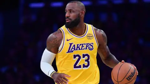 Reuters LeBron James dribbling a basketball up the court while looking to his right in close-up. He is wearing a yellow Los Angeles Lakers jersey with the number 23 written on the front. A white armlet is tied on his right arm.