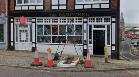 A Google image of a pub. It is a black and white painted building. Three traffic cones are in front of it.