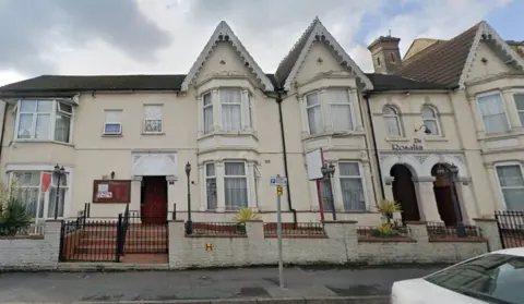Google Maps An off white building. There is a cloudy grey sky above it. There is the back of a white car to the right of shot.