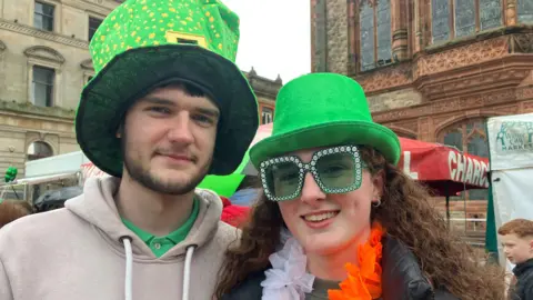 A man and woman are smiling. They're both wearing green hats. The woman has large green sunglasses on. 