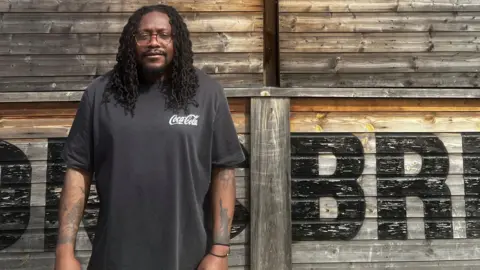 Gordy Mazvihwa A man with long curly black hair is wearing a black T-shirt. He has tattoos on his arms and wearing glasses. He is standing in front of a wooden building with the words "Dhillon's Brewery" in large black letters