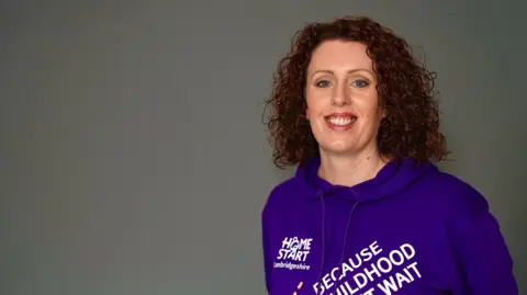 BBC/Martin Giles Woman with reddish dark shoulder length curly hair smiling to camera. She's wearing a purple hoodie with with Home-Start branding.