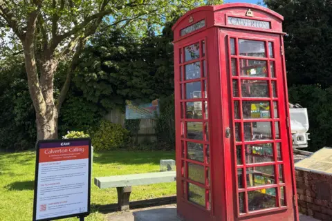 SUPPLIED Red phone box in front of green grass with a sign to the left of it