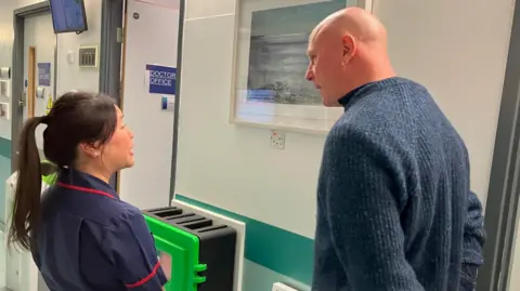 Ward sister Tamsyn Pattenden is talking to Kurt Jackson in the Phoenix ward while they stand in front of one of his paintings. She is wearing a navy uniform with a red trim and has long dark hair tied in a pony tail. He has his hands held behind his back and is listening to her. He is wearing a blue woollen jumper and has silver hoop earrings. The painting is a seascape in muted greys and blues.