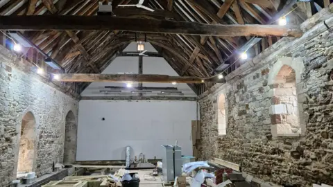 Poole Museum / BCP Council The interior of a medieval hall with stone walls and a pitched wooden roof. The floor is under construction and the area is covered with tools and components