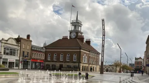 Stockton Town Hall. It is a stone and brick building with windows running alongside the front and sides. It has a clock positioned on its tower and a flag flying from a pole. The area in front has a display of fountains, while shops can also be seen along High Street.