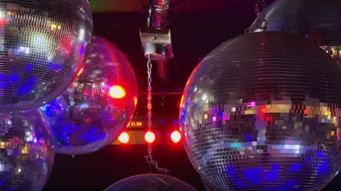 Image shows sparkling silver glitterballs hanging in a nightclub, with blue, pink and orange lights reflecting from them 