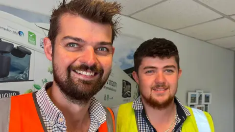 BBC Head and shoulders of Matt and Henry Bryan standing in an office on the farm. They both have dark hair and beards and are wearing checked shirts. Matt has a yellow high vis vest and Henry has an orange one