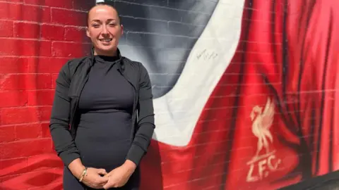 BBC/Angela Ferguson A woman with tied back dark hair is smiling while wearing black gym gear and standing next to a section of the main mural, with Sir Kenny's autograph on the white of his football shirt collar.