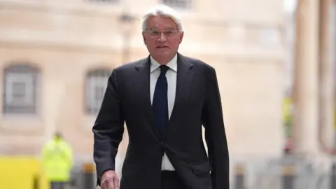An older man with short grey hair is pictured as he is walking away from a large stone building that is blurred out in the background. He is wearing a black suit, white shirt and blue tie. He is also wearing clear-framed glasses.