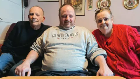 Mark Banks (middle) is sitting between is parents smiling at the camera.