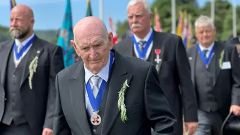 Cecil Raymond Gawne, with Paul Costain, Steven Carter and Peter Quayle walking behind him. The are all wearing dark suits and the Captain of the Parish Medals.