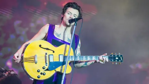 PA Media Harry Styles performing on the main stage during the BBC Radio 1's Big Weekend in 2022. He holds a yellow guitar and wears a purple jumpsuit