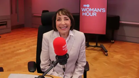 A woman with a bob haircut wearing a beige blazer sits in front of a red microphone.