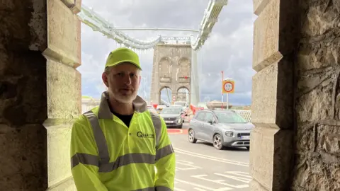 Dale Spridgeon A middle-aged man wearing yellow hi-vis on a large bridge crossing. There's cars travelling in both directions.