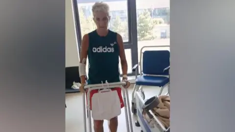 Carmel McConnellogue A woman using a zimmer frame to walk. She is wearing a navy vest top and has short grey hair.