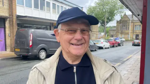 Gemma Dillon/BBC A gentleman is standing on a street with parked cars behind him.
He is wearing a dark blue cap, tinted glasses and a cream coat.
