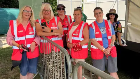 Cuffe & Taylor Six people, standing under an awning, smiling and looking at the camera. Five are wearing pink high-vis jackets. Two people to the left have a metal railing between them and are holding devices. Two people to the right have a number of red wristbands along their arms. They are standing on grass. 