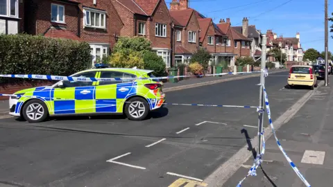 A police car is parked across a road. There are semi-detached houses in the background and blue and white police tape across the road in front of the car.