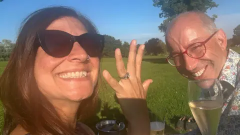 Outdoor photograph taken in a grassy park on a sunny day. Two adults sit on a picnic blanket with glasses of sparkling wine. The person in the foreground wears dark sunglasses and holds up a hand to display a silver ring on the ring finger. The second person, wearing red-framed glasses and a patterned shirt, sits opposite holding a drink. Trees and open green space are visible in the background beneath a clear blue sky.