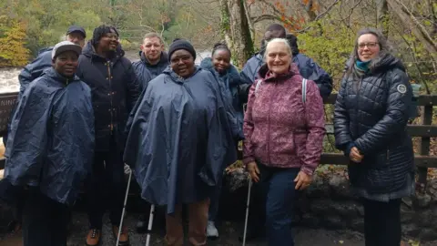 A mixed group of people stand in front of a fence by a river, posing for a picture. They all wear waterproof coats and winter clothing.