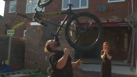 A man balances a bike on his chin outside a house as a woman laughs in the background