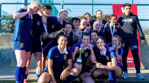 Fifteen members women's team wearing navy blue t-shirts and shorts and long socks pose on a pitch. A women at the front holds a gold cup and they all have medals around their neck, some holding them between their teeth.