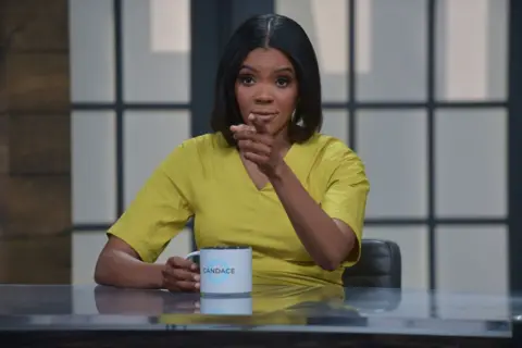 Getty Images Owens behind a desk holding a mug which says "Candace" on it, pointing towards the camera.