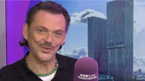 BBC Manchester Uma pessoa está sentada em um ambiente fechado em frente a um cenário que mostra dois altos arranha-céus modernos contra um céu parcialmente nublado. Em primeiro plano, há um microfone roxo com o texto “BBC RADIO MANCHESTER” impresso em branco. O indivíduo está vestindo um top escuro e um colar de contas verdes. O cenário parece ser um estúdio de rádio ou espaço de entrevistas.