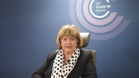 PA Baroness Heather Hallett sits in the inquiry chair wearing a black suit and spotty scarf, with the UK Covid-19 Inquiry logo on the wall behind her