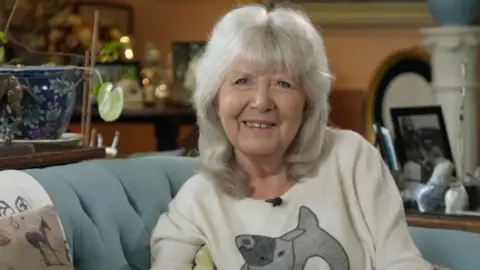 Jilly Cooper smiles at the camera. She's wearing a white jumper and sitting on a light blue sofa