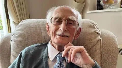 Man wearing glasses looks straight into the camera lens. He's smiling and wearing a white shirt with a blue and white spotted tie and grey cardigan