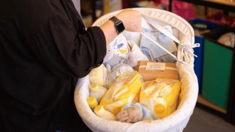 A person in a black jumper holding a basket filled with baby suplies, such as nappies and toys.