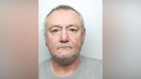 The mugshot of David Jack, a man with light grey hair and a light moustache with stubble. He is starting impassively forwards. 