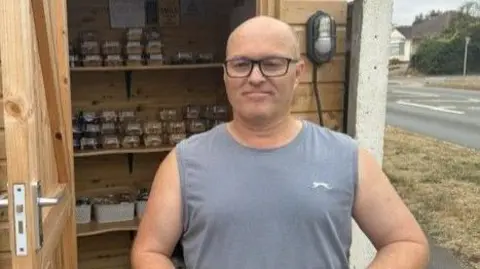 Julian Manser stands outside the Capel Cake Shed with shelves of packaged cakes visible behind him.