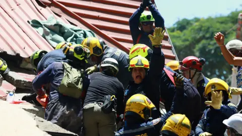 Reuters Firefighters react as they rescue a person following an earthquake, in Cali, Colombia, August 11, 2026 - one can be seen prominently in the middle, holding his arm up and appearing to shout 