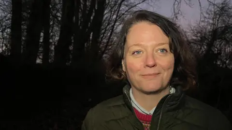 Eliza Whitaker standing in front of trees, looking past the camera. It appears to be dusk