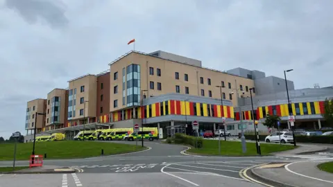 A hospital with ambulances parked outside it. The hospital building is several storeys high, and has red and yellow cladding on the lower part.