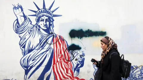 An Iranian woman in a leopard print headscarf walks past an anti-US mural on a street in Tehran, showing a the Statue of Liberty in red and white with damage to its arm. 