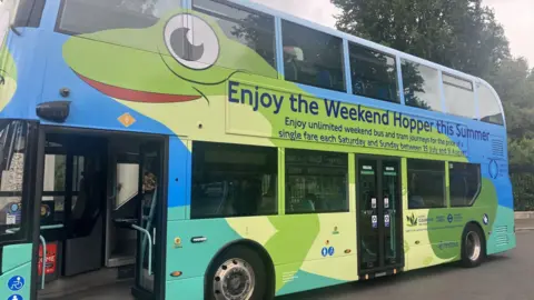 Side view of a London bus with a big advert for the Weekend Hopper bus service across its side featuring blue, green and yellow strands of colour and dark blue text