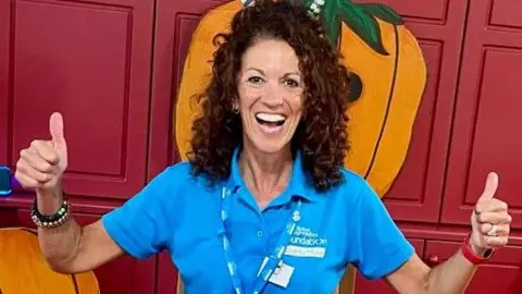 Jules Jenkinson, a woman with dark and curly hair, is pictured grinning widely at the camera and raising both of her thumbs. She is wearing a blue polo shirt. 