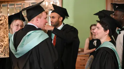A group of people making small talk. They are wearing black graduation caps and black gowns with a green hood. 